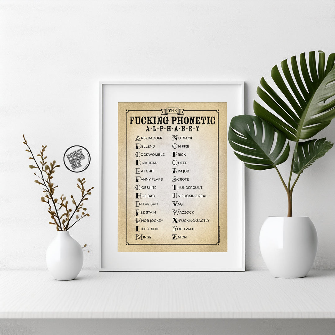 The "Rather Rude and Funny Fucking Phonetic Alphabet Print" is framed on a white shelf between two white vases with green leaves and dried branches—perfect funny home office or phonetic alphabet wall art. A small round pin sits nearby.