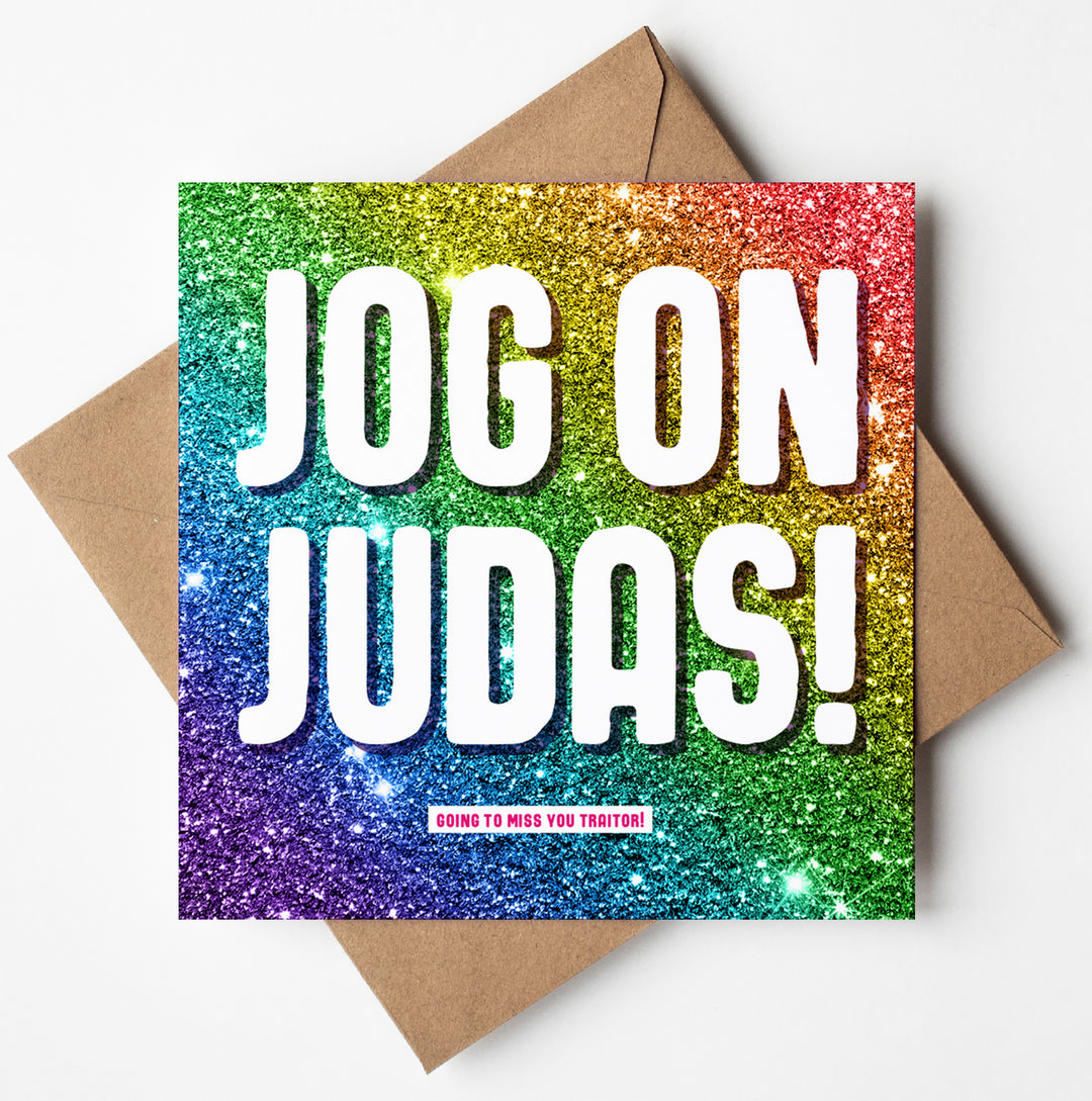 The "Jog On Judas Glitter Card" showcases a humorously cheeky farewell message against a sparkling rainbow backdrop, with prominent white text exclaiming "JOG ON JUDAS!" and smaller text below saying "GOING TO MISS YOU TRAITOR!" It is paired with a brown envelope, making it an ideal choice for a lighthearted retirement send-off.