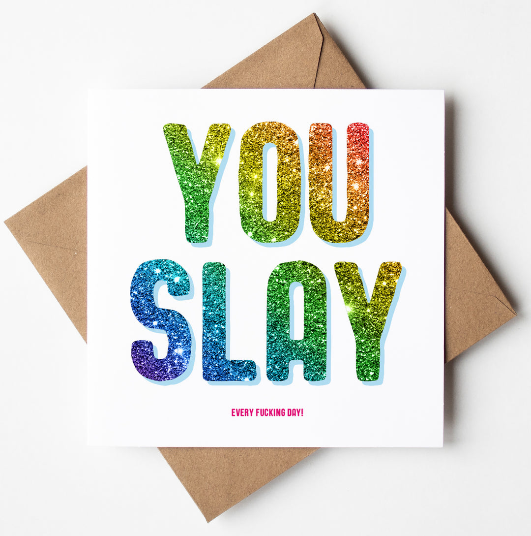 The "You Slay Every Day" card features a glittery, rainbow-colored design with the bold statement "YOU SLAY" on a clean white background. Below, in smaller text, it exclaims "EVERY FKING DAY!" The card comes presented in a brown envelope and is shipped worldwide in a biodegradable bag for an eco-friendly experience.