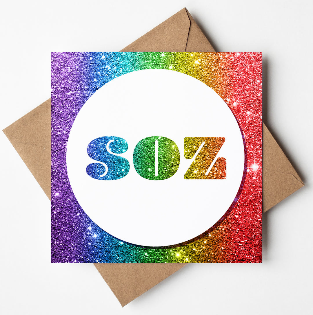 Product Description: The Soz Rainbow Glitter Card is a square card with "SOZ" boldly displayed in iridescent letters inside a white circle, bordered by a vibrant, sparkling rainbow glitter frame, all beautifully paired with a brown envelope.