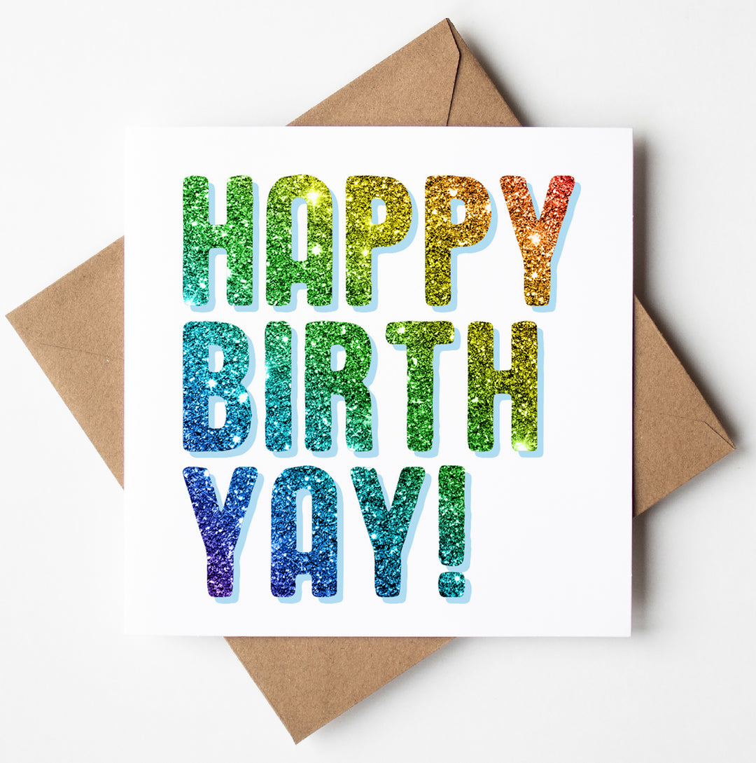 A typographic birthday card enclosed in a brown envelope, featuring the holographic text "Happy Birth Yay!" in shimmering rainbow colors on a clean white background.