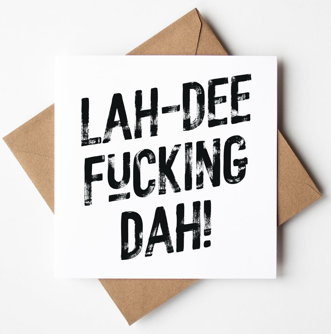 The Lah-Dee Fucking Dah! Harlem Card features bold, distressed black typography proclaiming "LAH-DEE FUCKING DAH!" and comes with a brown envelope, all made using biodegradable packaging. The card is centered against a white background.