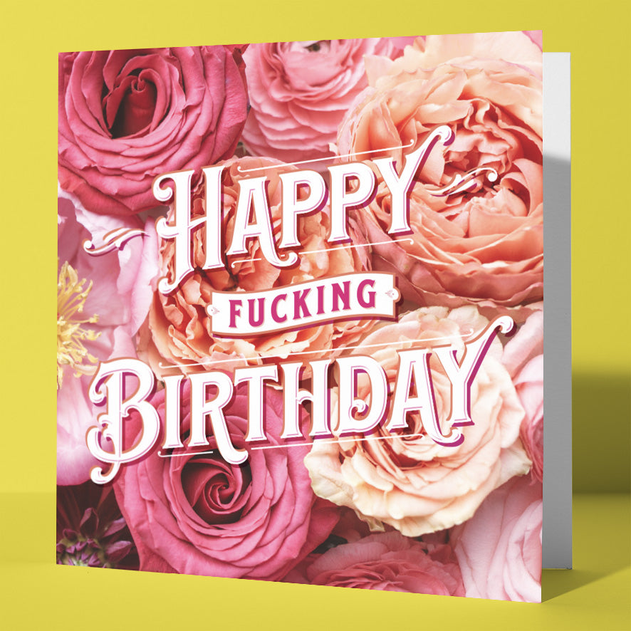 The Happy Fucking Birthday Ranunculus Card features pink and peach roses in the background with bold white and pink text. Designed in Wales, it stands out against a yellow surface—perfect for a cheeky birthday greeting.