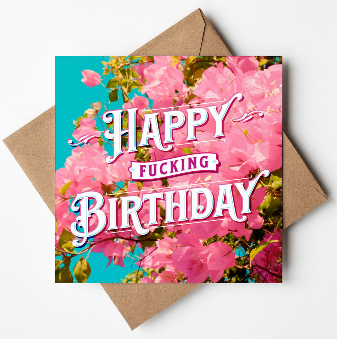 A square card designed in Wales, called the Happy Fucking Birthday Bougainvillea Card, showcases pink flowers and vibrant green leaves against a turquoise background. It features bold white and pink text that says "Happy Fucking Birthday" and comes with a brown envelope.