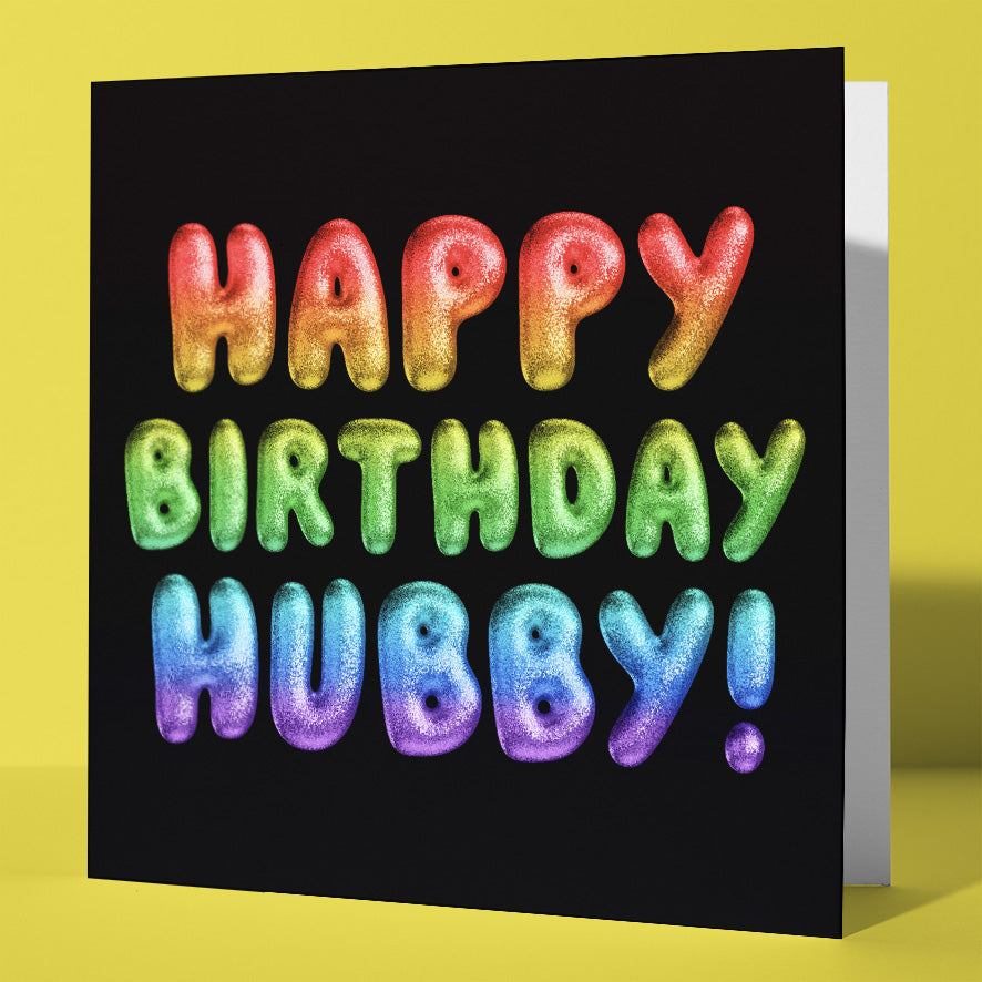 The Happy Birthday Hubby! Rainbow Font Card features colorful, puffy letters on a black background and stands out against a yellow surface. Designed in Wales, it's the perfect vibrant touch for any birthday celebration.