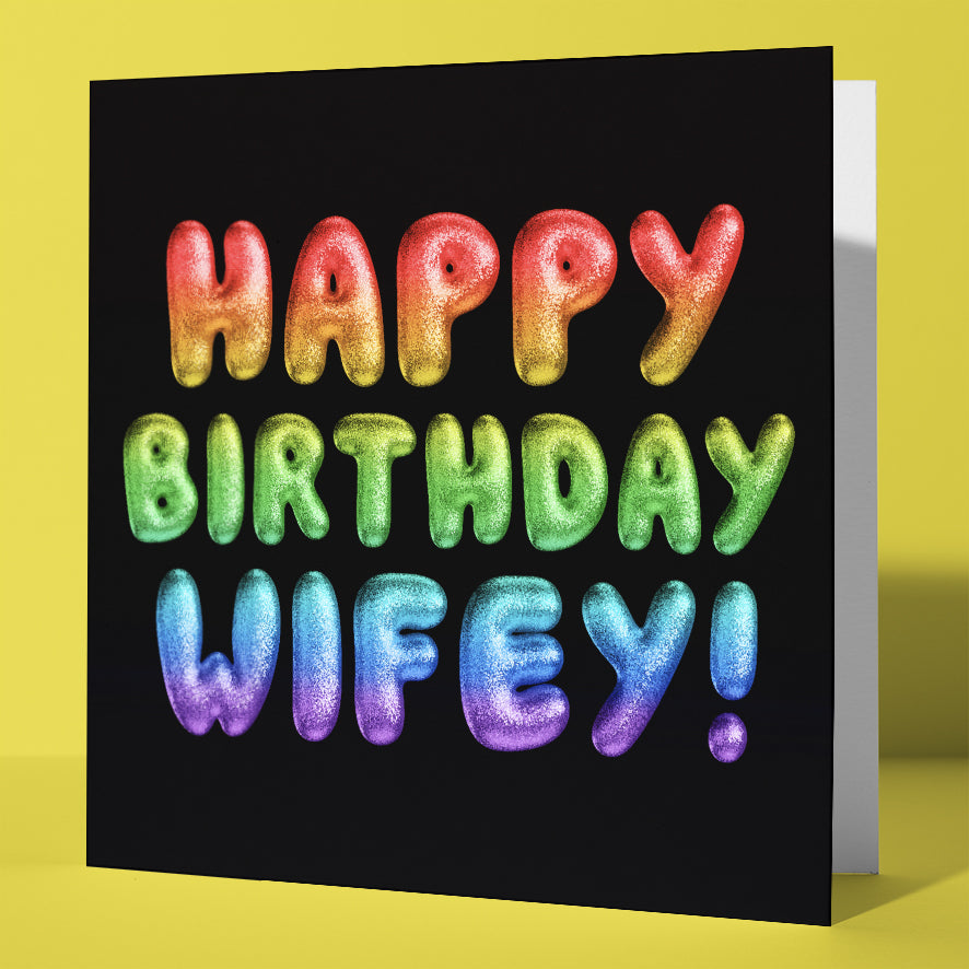 The Happy Birthday Wifey! Rainbow Font Card features a glittery rainbow font on a black background. Standing upright on a yellow surface, it’s perfect for her special day. UK shipping available.