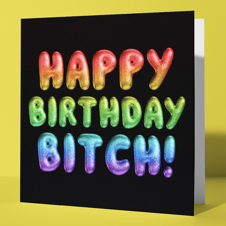 The Happy Birthday Bitch! Rainbow Font Card features glittery balloon letters on a bold black background and stands upright on a yellow surface, making it the perfect statement piece for any celebration.