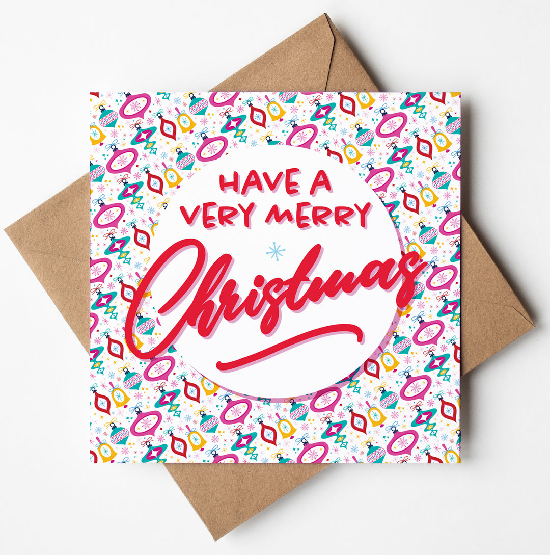 Introducing the "Have a Very Merry Christmas Bright Baubles Card"! This festive greeting card features the message "Have a Very Merry Christmas" elegantly displayed in red and pink on a white circular area. The vibrant background showcases abstract patterns in shades of pink, orange, and teal. The card is blank inside, allowing you to add your personal touch, and it comes with a brown envelope packaged in biodegradable material.