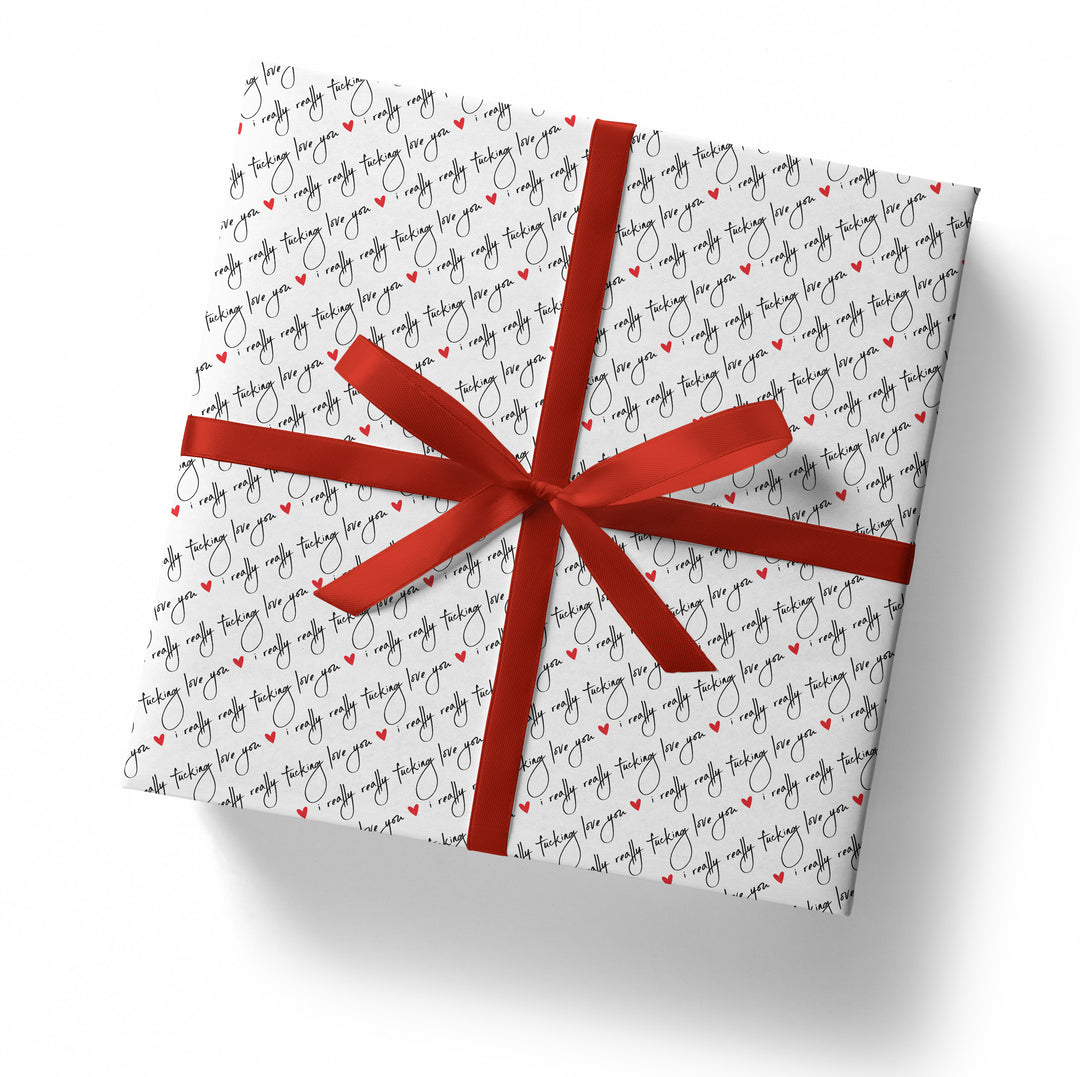 A square gift box wrapped in "I Really Really Fucking Love You Gift Wrap"—white paper with black cursive text and red hearts—finished with a wide red ribbon and bow, sits on a white surface.