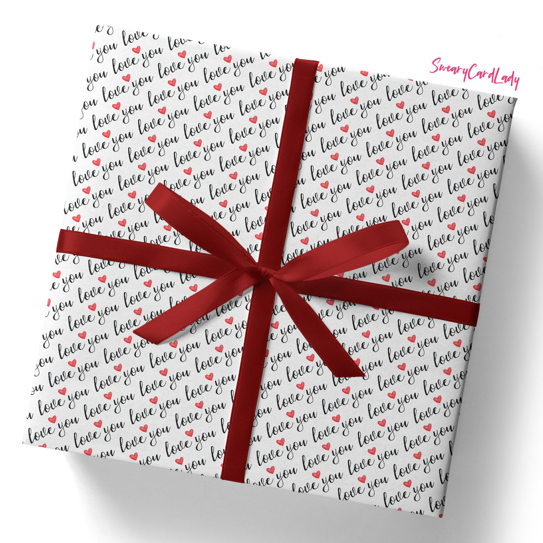 A square gift box wrapped in Love You Gift Wrap (Folded Sheet) featuring black and red "love you" text with red hearts, tied with a shiny red ribbon. "SnazzyCardLady" appears in pink—ideal for anniversary gifts.