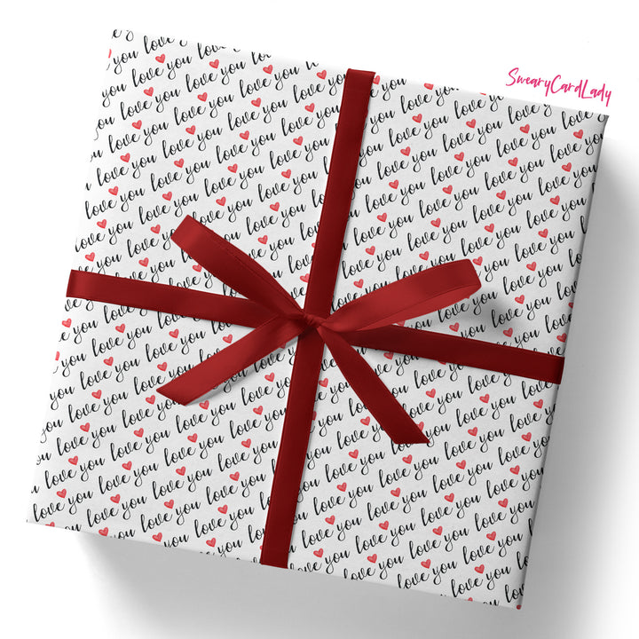 A square gift box wrapped in Love You Gift Wrap (Folded Sheet) featuring black and red "love you" text with red hearts, tied with a shiny red ribbon. "SnazzyCardLady" appears in pink—ideal for anniversary gifts.