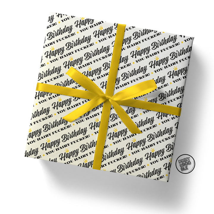 A folded sheet from Prints Doodle, named the Hairy Fucker Birthday Gift Wrap, is wrapped around a gift box. The funny white paper features repeating black and gold text that reads "Happy Birthday, You [Expletive]," and is tied with a yellow ribbon bow. A circular tag in the bottom right corner has a faint smiley face—making it an ideal humorous birthday gift for a husband.