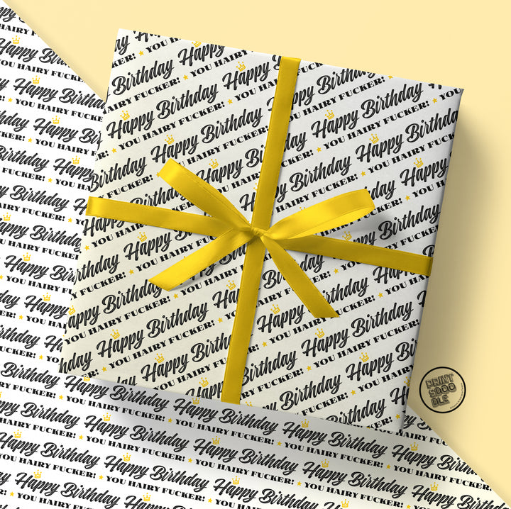 A gift wrapped in Prints Doodle's Hairy Fucker Birthday Gift Wrap (Folded Sheet), featuring the playful "Happy Birthday You Fucking Fuck" phrase in various fonts and colors. Adorned with a yellow ribbon and bow, this makes the perfect birthday gift for your husband. The background showcases additional wrapping paper with the same bold design.