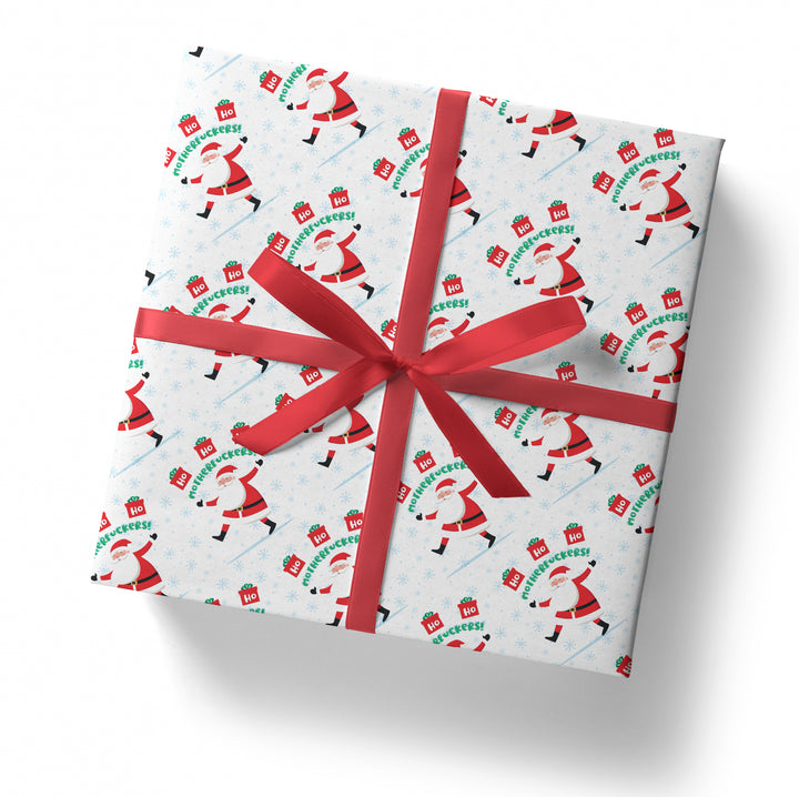 A square gift box wrapped in "SALE! Ho Ho Ho Motherfuckers!" folded sheet, decorated with a red ribbon and bow, featuring dancing Santa Claus and "Ho Ho Ho" signs, viewed from above on a white background.