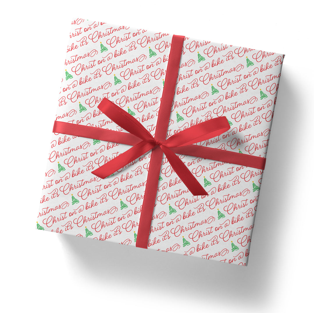A gift box wrapped in the SALE! Funny 'Christ on a Bike It's Christmas' Gift Wrap (folded sheet), featuring red festive text and green trees, tied with a red ribbon and bow—perfect for secret Santa gifts, shown from above on a white background.