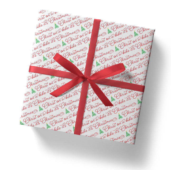 A gift box wrapped in the SALE! Funny 'Christ on a Bike It's Christmas' Gift Wrap (folded sheet), featuring red festive text and green trees, tied with a red ribbon and bow—perfect for secret Santa gifts, shown from above on a white background.