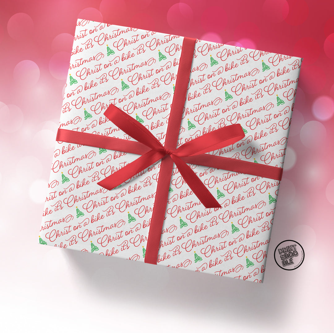 A square gift box wrapped in SALE! Funny 'Christ on a Bike It's Christmas' Gift Wrap (Folded Sheet), tied with a red ribbon, sits against a pink and white bokeh background.
