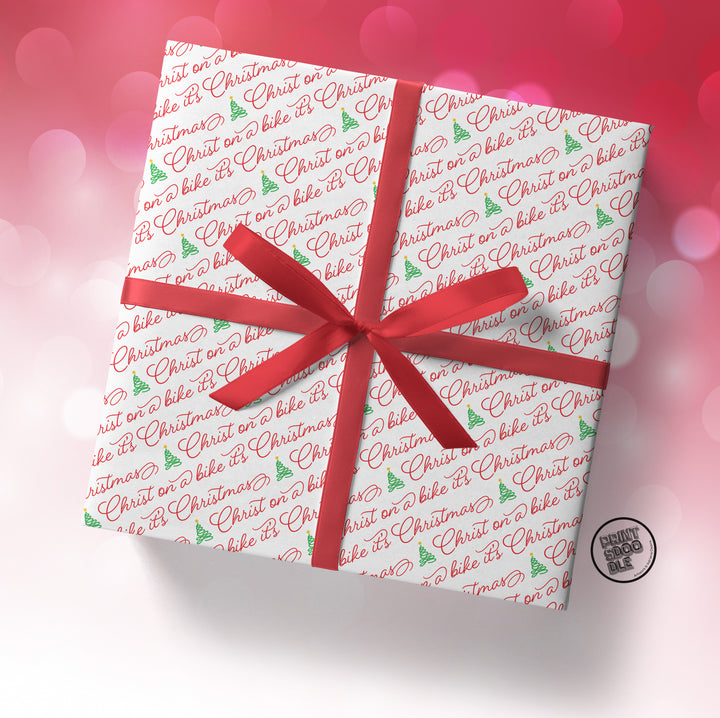A square gift box wrapped in SALE! Funny 'Christ on a Bike It's Christmas' Gift Wrap (Folded Sheet), tied with a red ribbon, sits against a pink and white bokeh background.