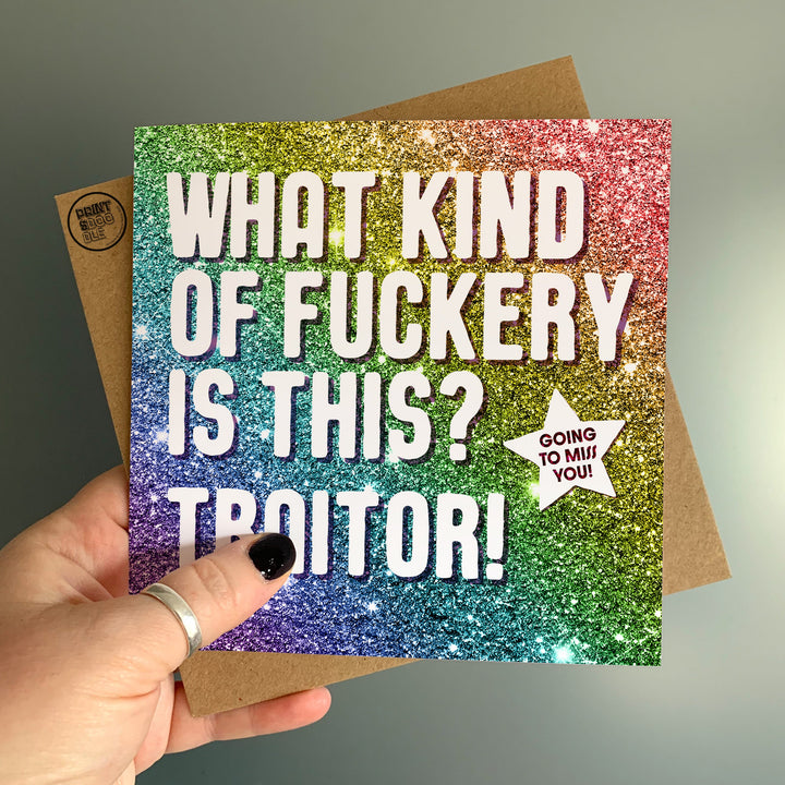 A hand holds the "What Kind Of Fuckery Glitter Card," showcasing a glittery rainbow background with the text, "WHAT KIND OF FUCKERY IS THIS? TRAITOR!" and a small starburst that reads, "GOING TO MISS YOU!" It's an ideal cheeky farewell card for someone moving away. A brown envelope peeks out from behind the card.