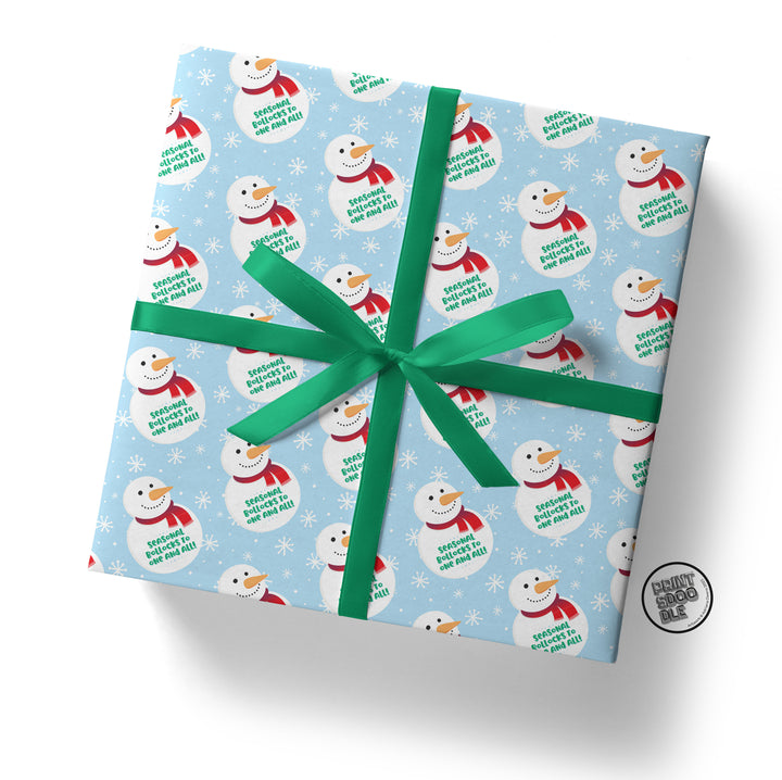 A Secret Santa gift wrapped in Seasonal Bollocks Christmas Gift Wrap, featuring cheerful snowmen sporting red scarves and black hats, all with smiling faces, on light blue paper with green text. It is tied with a green ribbon and adorned with a circular sticker in the corner that reads "DEADLY NIGHT SHADE.