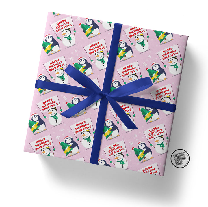 A square gift box wrapped in "Happy Birthday Baby Jesus" gift wrap, featuring cute snowman illustrations and adorable penguins with the text "Happy Christmas, Best Wishes." A cheerful blue ribbon ties it all together, enhancing its festive charm.