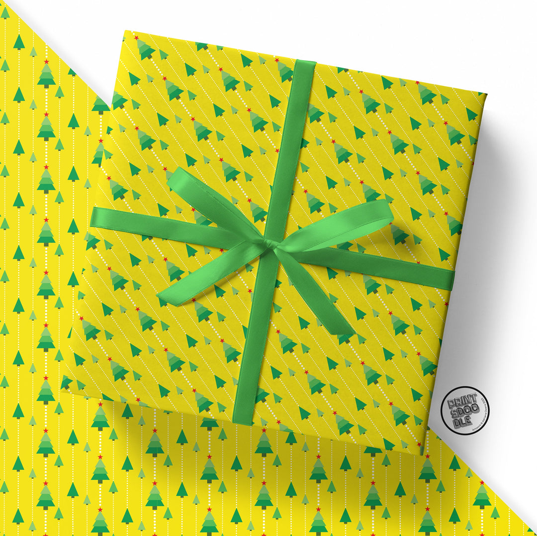 A gift is wrapped in a vibrant yellow folded sheet featuring a funky Christmas tree pattern, tied with a green ribbon, and placed on matching wrapping paper. A circular logo, partially visible, reads "Infinity Book Box" in the corner.